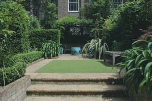 A small, neatly landscaped garden with stone steps, a rectangular patch of artificial grass, green plants that attract bees, and a table with chairs in the background.