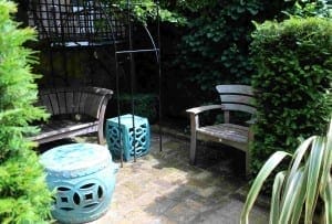 Two wooden chairs and two blue ceramic stools are arranged on a brick patio in a small, leafy garden area with green hedges, plants, and pyramids of vibrant greenery for a tranquil outdoor retreat.