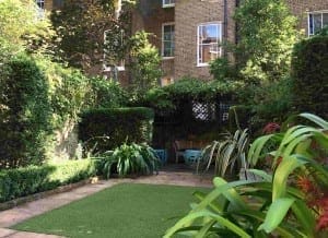 A small urban garden with a paved patio, green plants, hedges, and two blue chairs by a wooden table against a brick building—its geometric layout subtly echoes the orderly lines of ancient pyramids.