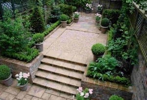 A neatly landscaped garden with stone steps, potted plants, trimmed shrubs, and flower beds forms a tranquil space. In the center, a gravel area features small pyramids, all enclosed by brick walls and green fencing.