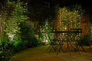 A metal patio table with four chairs sits on a stone terrace at night, surrounded by lush plants and illuminated by string lights on a garden trellis, creating a Trompe l’Oeill effect of depth and enchantment.