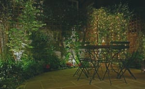 A patio at night with a round metal table and four chairs, surrounded by greenery and illuminated by string lights and garden lighting, features Trompe l’Oeill accents that enhance the enchanting atmosphere.