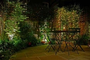 A small outdoor patio with a round table and four chairs, surrounded by plants and illuminated by string lights and garden lighting at night, features Trompe l’Oeil art that creates the illusion of an even larger, enchanting garden space.