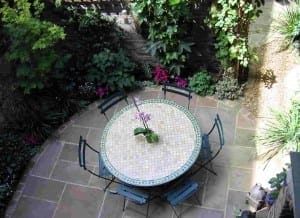 A round mosaic table with four chairs sits on a stone patio, surrounded by green plants and flowers in a garden setting. At the center, an orchid in a pot adds charm, while Trompe l’Oeill details enhance the enchanting scene.