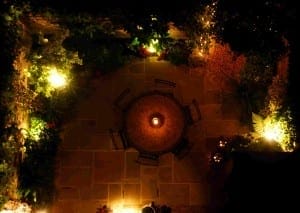 A round table with chairs is centered on a stone patio, surrounded by plants and illuminated by warm outdoor lighting at night, with Trompe l’Oeill details enhancing the enchanting atmosphere.