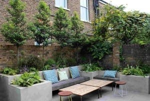 Outdoor patio with concrete benches, patterned cushions, small wooden tables, and lush greenery against a brick wall creates a bespoke garden setting.
