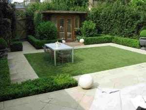 A modern garden designed as one of the ultimate outdoor rooms, featuring a rectangular artificial lawn, a white table and chairs, neatly trimmed hedges, stone paving, and a wooden garden shed with glass doors.