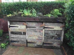 A three-bin compost binshed system made from reclaimed wood, with plants growing on top, is situated in a garden with a hedge in the background.