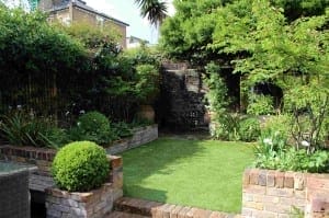 A small, well-maintained garden with brick planters, green shrubs, a fake lawn, and a wooden bench near the back fence.