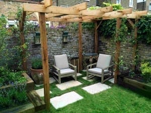 Two cushioned chairs sit under a wooden pergola with sturdy beams in a small garden, featuring a stone path, potted plants, and a brick wall in the background.