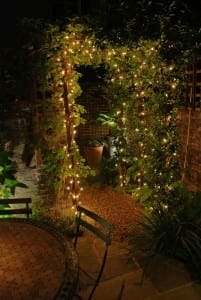A garden archway covered in greenery is illuminated by outdoor lighting and string lights at night, with a patio table and chairs in the foreground.