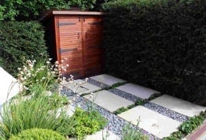 A wooden garden shed and binshed stand beside tall hedges, with a path of large square stone tiles and gravel leading to them. Green plants and white flowers border the walkway.