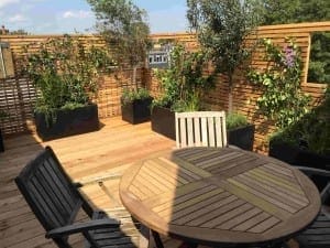 Wooden patio with a round table, four chairs, and planter boxes filled with greenery offers a framed view through a wooden slat privacy fence on a sunny day.