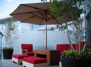Outdoor patio on a roof garden featuring red cushioned chairs, a wooden table, potted plants, and a large brown umbrella set on a tiled floor.