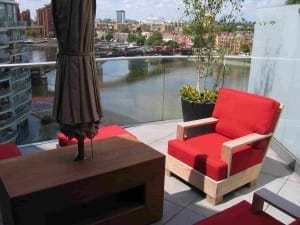 A balcony designed like a cozy roof garden, featuring red cushioned chairs, a table, a closed umbrella, potted plants, and views of the river and city buildings in the background.