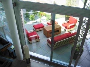 Outdoor patio area viewed through glass doors, featuring wooden furniture with red cushions arranged around a square wooden table in a charming roof garden setting.