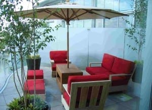 Outdoor patio seating area with red-cushioned chairs, a wooden table, potted plants, and a large beige umbrella on a tiled roof garden.