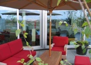 Outdoor patio with red cushioned chairs and wooden table under a large umbrella, set near glass doors with reflections, surrounded by potted plants—perfect for enjoying a relaxing moment in this inviting roof garden.
