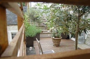 Wooden deck patio with potted plants, a table, two chairs, and a mug, viewed through a wooden railing. Steps and greenery evoke the calm of a Mondrian Garden in the background.
