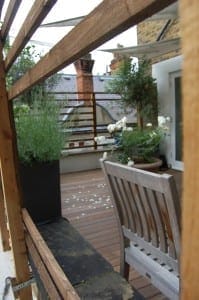 A wooden patio with potted plants, a wooden bench, and a Mondrian Garden style view of rooftops and chimneys in the background.