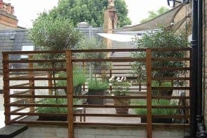 A rooftop patio, known as the Mondrian Garden, features potted plants and small trees, enclosed by a wooden slat fence, with buildings and trees visible in the background.