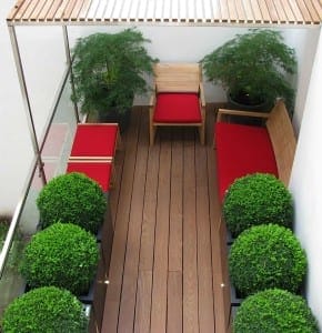 A small balcony with wooden flooring, red-cushioned chairs and bench, potted green plants including graceful Acers, and a wooden slatted canopy overhead.