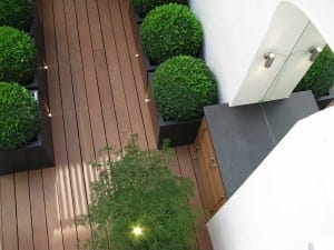 Overhead view of a modern patio with wooden decking, round potted acers and shrubs, a square stone countertop, and a white wall with an arched mirror.