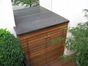 A wooden outdoor cabinet with horizontal slats and double doors, topped with a dark stone countertop, stands beside green acers and a white wall.
