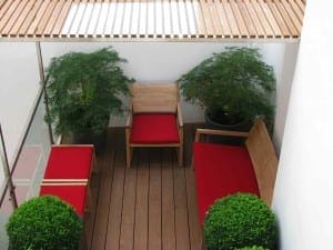 Small patio area with wooden seating featuring red cushions, two side tables, potted green plants including vibrant acers, and a wooden slatted overhead cover.