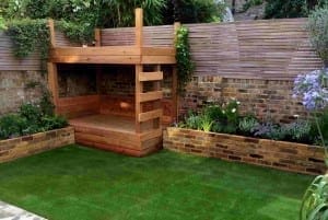 A small backyard with artificial grass, raised brick planters, pergolas beams above a wooden play structure with a ladder in the corner—all surrounded by a wooden fence and lush plants.