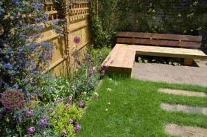 A bespoke garden bench with a backrest sits in a corner of a garden with green grass, flowering plants, and a wooden fence covered in climbing plants.