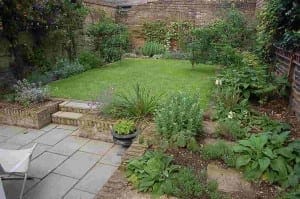 A small walled garden with a rectangular lawn, surrounding flower beds, brick steps, and a paved patio area with a planter and white chair—perfect for relaxing or starting your own Edible Gardens adventure.