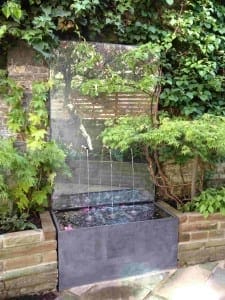A vertical glass water feature with streams of water flowing into a rectangular basin, surrounded by green plants and brickwork, brings modern elegance to garden water features.