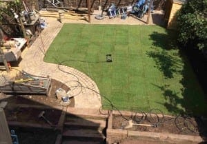 A backyard in Queens Park with newly laid turf, a curved stone pathway, scattered garden tools, and construction materials around the edges.