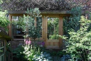 A wooden garden shed with glass doors and windows creates inviting outdoor rooms, nestled among dense green plants and flowers, with branches arranged on the roof.
