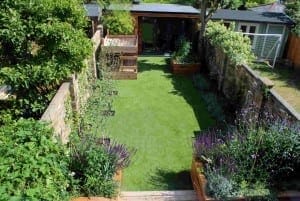 A rectangular backyard features fake lawns, raised wooden planters, lush green shrubs, and a small outbuilding tucked at the far end.