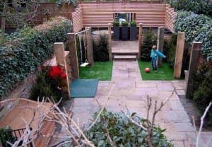 A child in a blue jacket plays with a red ball in a backyard garden featuring artificial grass, a swing set, and a patio area.
