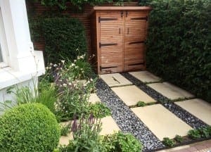 A small garden features large pavers with stone textures and gravel strips between them, bordered by green plants and shrubs, with a wooden storage shed at the back.
