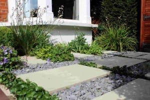 A modern garden path with stone textures featuring rectangular paving stones and gray gravel, surrounded by various green plants and flowers, beside a house exterior.