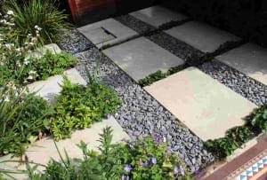 Rectangular paving stones with natural stone textures are laid in a grid pattern with gray gravel in between, bordered by green plants and flowers along the garden pathway.