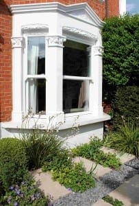 A white bay window with decorative trim and subtle stone textures on a brick house, surrounded by green shrubs and flowering plants in a landscaped front garden.