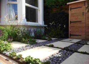 Small garden with stone textures on slabs and gray gravel paths, bordered by flowering plants and greenery, next to a wooden shed and a white house window.