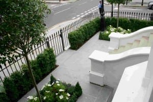 A small, neatly landscaped Italianate garden with a paved walkway, trimmed hedges, flower beds, black metal fence, and a street visible in the background.