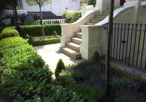 Stone steps lead up to a white building entrance, surrounded by neatly trimmed hedges and lavender plants in the style of an Italianate Garden, all enclosed by a sleek black metal fence.