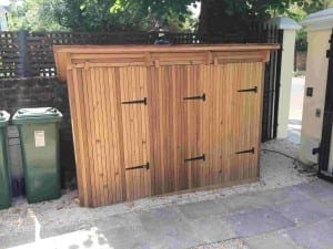 A wooden Binshed with three doors and black metal hinges, placed on gravel beside two green bins and a wooden fence.