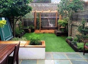 Small urban garden with fake lawns, a wooden pergola, raised plant beds, a patio dining area, and trees all bordered by classic brick walls.