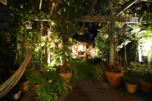 A well-lit garden at night with outdoor lighting highlighting potted plants, a hammock on the left, and a small illuminated structure at the far end.