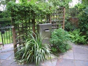 Metal mailbox surrounded by green plants and shrubs, with water features nearby, set against wooden lattice fencing in a garden with stone paving.
