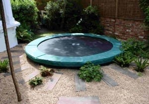 An in-ground trampoline with a green safety pad is nestled in a garden area, surrounded by gravel, wooden planks, lush plants, and vibrant gardens.