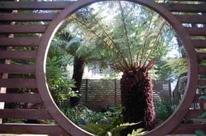 A circular wooden window frames a view of a garden with ferns and greenery, where sunlight streams through trees and pergolas beams overhead.